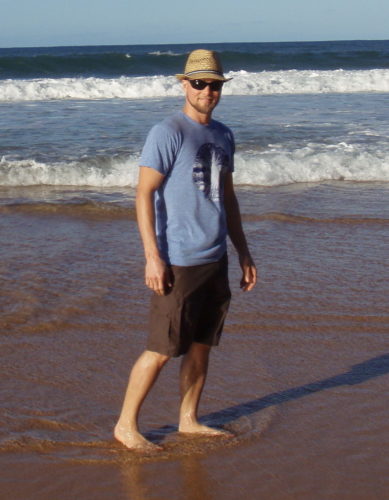 Scott, as an adult, standing barefoot on the beach. There's a thin layer of water under his bare feet and waves in the background. He's wearing a straw hat, sunglasses, a light blue short sleeved tshirt and brown knee length cargo shorts