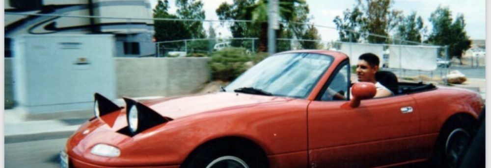 Rudy driving a red Miata. The background is blurry.