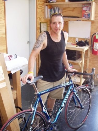 Robin with his Trek bike. He's wearing a black tank top and yellow sunglasses. He has a large tattoo on his right shoulder and a yellow LIVESTRONG band on his wrist. He's standing in front of a white door with a silver knob and light wood shelves and work tables in what appears to be the corner of a garage