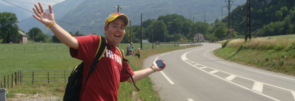 Jimmy standing by the side of the highway at the Tour de France. He is carrying a backpack and holding his cell phone in his left hand. His arms are out wide and he's standing on his left foot. He's wearing a yellow LIVESTRONG baseball hat and a red Stanford t-shirt with Stanford written in white on the front.
