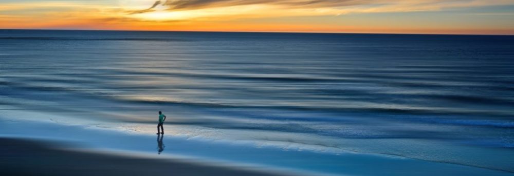 The ocean at sunset. There's the silhouette of a man walking on the left. The water is varying shades of blue. The sky is cloudy with some orange.