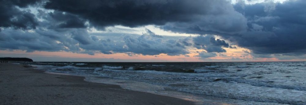 A gray, cloudy day at the ocean. On the left in the front, a triangle of sand is visible. The large cloud in the foreground is dark gray; the one behind is light gray.