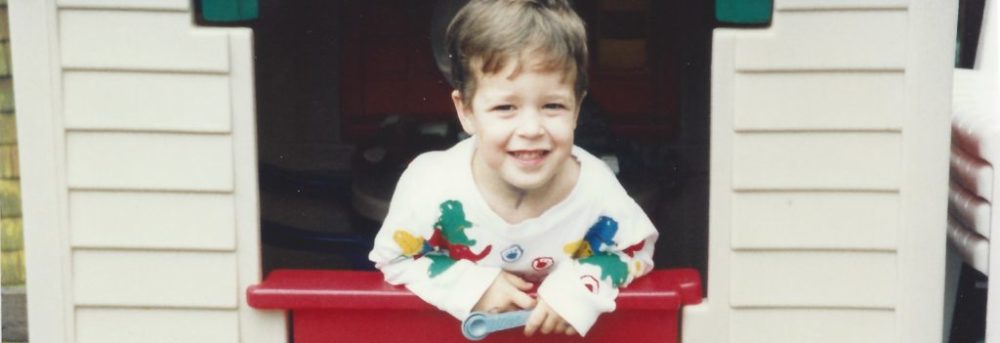 Jimmy, wearing a white long sleeve t-shirt, with different primary color dinosaurs on it. The playhouse has a green roof, red door and tan walls.