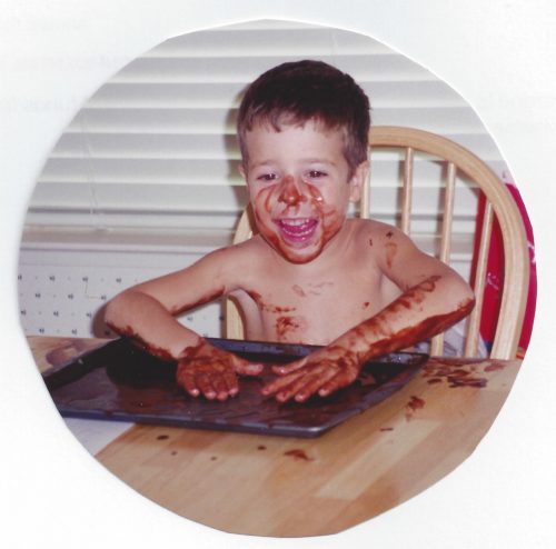 Jimmy, shirtless, sitting at the kitchen table with his hands flat in a cookie sheet with pudding in it. There's pudding on his hands, arms and face, and he is laughing.