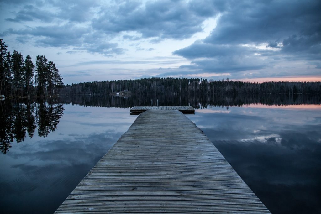 Wooden dock stretching into the distance. Beyond it is a row of trees. Water is on both sides. The sky is cloudy. and dark. The sun has set in the right of the picture leaving a faint bit of orange above the trees and reflected on the water.