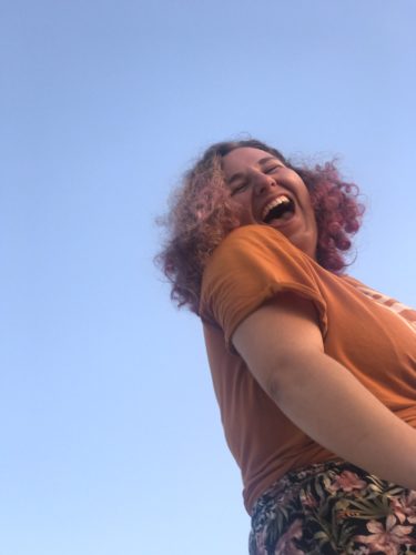 Eve leaning into the picture from the right. She's wearing a short sleeve rust colored t-shirt. She's laughing with her mouth open. Her light brown hair is shoulder length and parted on the left. Behind her is blue sky.