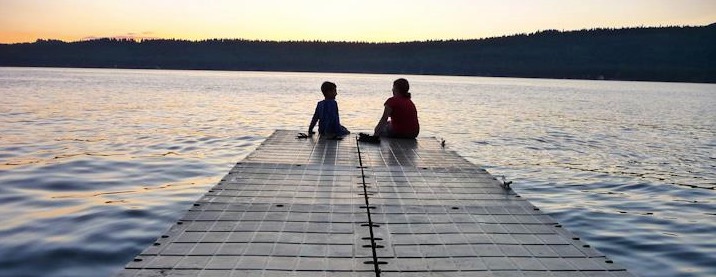 Eve and a younger sibling sitting at the end of a dock as the sun begins to set. The sky is light yellow with blue water between it and the kids