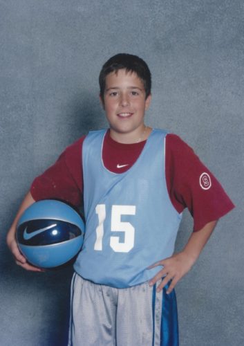 Jimmy wearing a pale blue basketball jersey with a white number 15 on it and a cardinal red Stanford t-shirt underneath. His hair is short and brown. He has a pale blue and black Nike basketball under his right arm. 