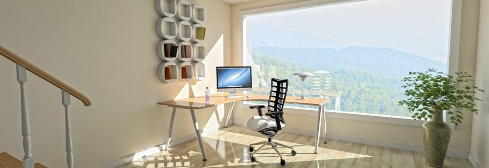 Modern desk with tan wood and white legs shaped to fit in the far corner of the room. There's a computer monitor on the desk and a black chair in front of the desk. The window runs the length of the wall and looks out on the mountains.
