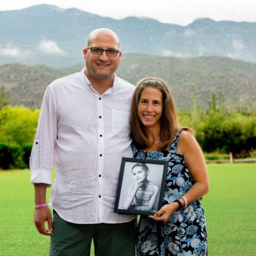 Erica and her husband standing on green grass in front of two hills, with wispy clouds in front of the higher one. Ariella's dad is wearing a white button down shirt with black buttons and jeans. He's bald with a close cropped beard and glass with black rims. Erica is holding a black and white photo of Ariella in a black frame and a sleeveless blue print dress on a black background. They're both wearing wristbands