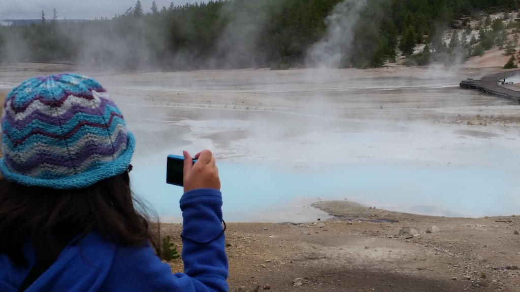 Clara taking a picture at Yellowstone of a geothermal pool, pale blue in color with white steam rising from it. Clara is wearing a royal blue long sleeve jacket and a knit cap with v shaped stripes of turquoise, light purple, white and deep purple