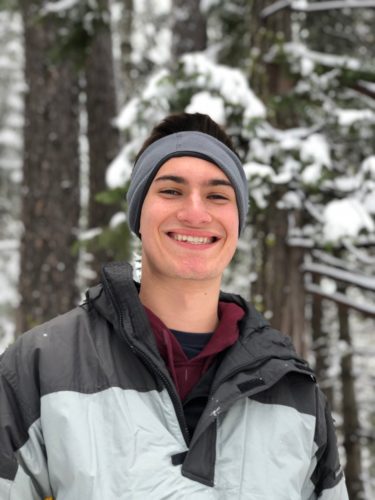 Brenden wearing a gray and black ski cap with a white pom pom, a maroon shirt and a dark gray and light gray ski jacket. Behind him are pine trees with snow on them