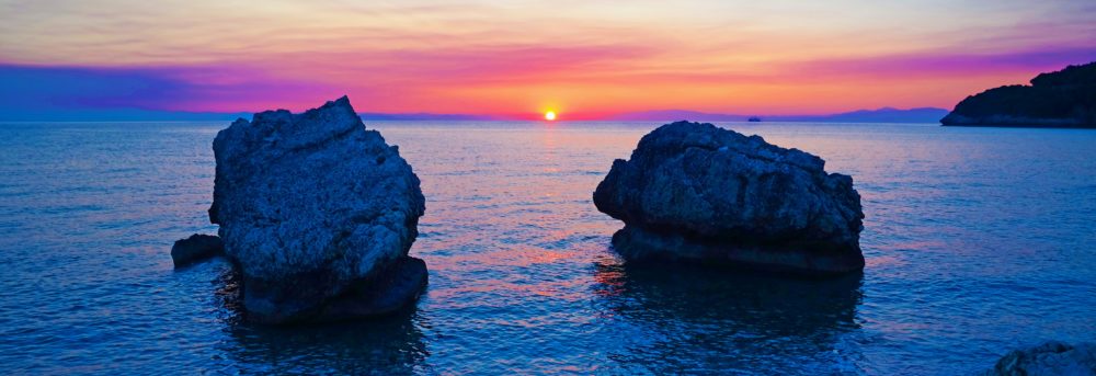Two large boulders in the ocean close to the shoreline. The sun is just above the horizon casting a pink and blue cast on the water and an orange band in the sky with blue above it