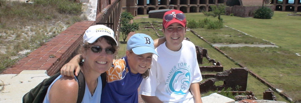 Margo, Molly, Jimmy sitting on a low stone wall in front of a fort in Florida. Margo is wearing a light blue tank top, white LIVESTRONG hat, sunglasses and wearing a backback. Molly is wearing a blue swim shirt with orange and white sleeve and blue basketball shorts. Jimmy is wearing a red Stanford hat with sunglasses perched on the brim, a white t-shirt with turquoise writing and black basketball shorts