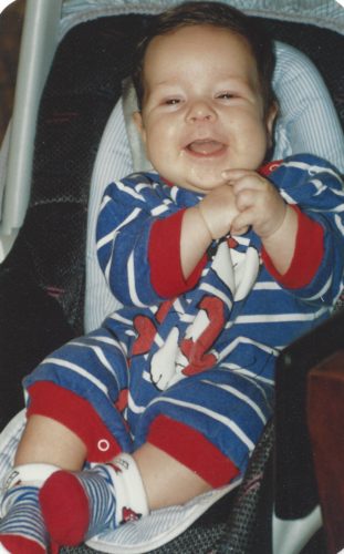 Baby Jimmy sitting in a carseat with a blue and white strip onesie with Snoopy dancing on the front and red cuffs on the arms and legs