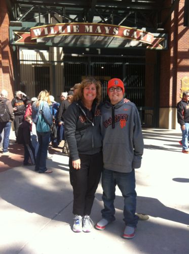 Margo and Jimmy standing outside of the Willie Mays gate at AT&T Park. Margo's wearing black pants and a gray zip up sweatshirt. Jimmy's wearing an orange beanie with SF on the front, glasses, jeans and a light gray sweatshirt with SF Giants across the chest in orange