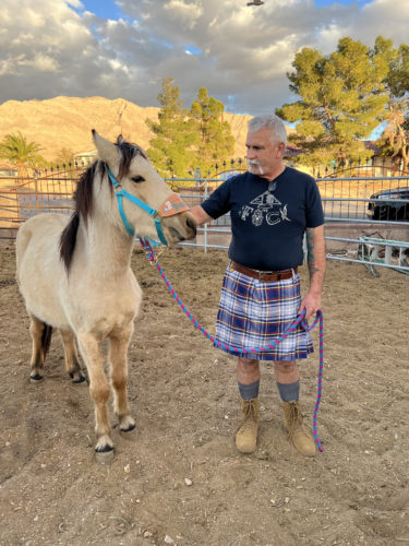 Max is wearing a red, black and white kilt and black t-shirt. He's standing next to a buckskin donkey wearing a teal halter