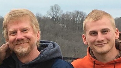 Roger and his son TJ. Roger is wearing a black hoodie. TJ is wearing an orange hoodie pullover. They both have blond hair. Roger has a blond mustache and beard