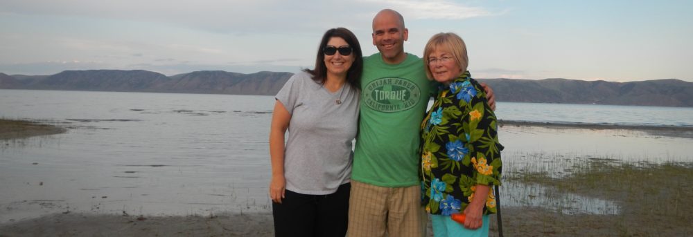 John, his wife Kathleen standing to his right and his mother standing to his left. Kathleen is wearing a gray t-shirt and black shorts. John is wearing a green tshirt and tan shorts. John's mother is wearing a yellow, green and blue print blouse with a black background and teal pants. They're standing on the shore of a body of water