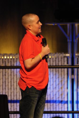 Jimmy on stage at the LIVESTRONG Challenge in 2010. He's holding a microphone in his right hand, wearing a short-sleeved red collared shirt and black pants. He has a LIVESTRONG band on his right wrist, and he's bald. 