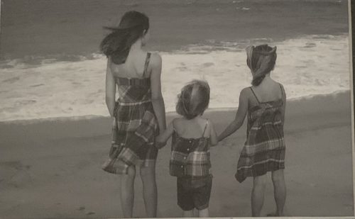 All three sisters looking at the ocean. They are each wearing dresses made of the same plaid material. The photo is in black and white