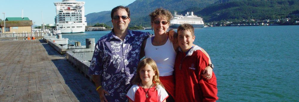 Dan, Margo, Molly and Jimmy in Alaska standing on the dock in front of a cruise ship. Dan is wearing a blue and white Hawaiian print shirt and sunglasses. Margo has her hair in a pony tail and is wearing a white tank top. Molly is wearing a red shirt with white sleeves and blue jeans. Jimmy is wearing a red jacket with a hood and jeans