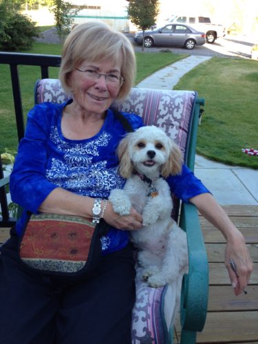 John's mom with a white poodle mix. She is sitting with her purse on her lab wearing a print shirt and a blue ¾ length sleeve jacket. She has blond hair and is wearing glasses