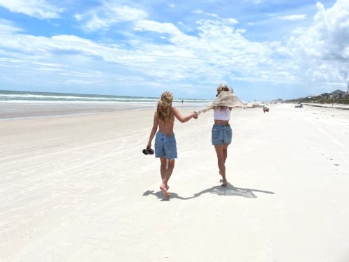 Tressa's two surviving daughters are walking on a sandy beach. Their backs are to the camera. The girl on the left is waring jeans shorts and carrying her shoes. The other daughter is wearing a white top, jeans shorts and a white shirt that's billowing up