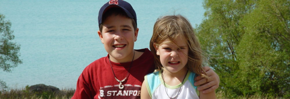 Jimmy and Molly sitting in front of a turquoise blue lake in New Zealand. Jimmy is wearing a black baseball cap and red Stanford tshirt. Molly is wearing a pendant and short sleeve white t-shirt with some splotches of light turquoise