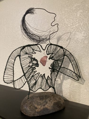 Wire sculpture of a person in mourning. Head raised, hands on the heart with a red heart in the middle