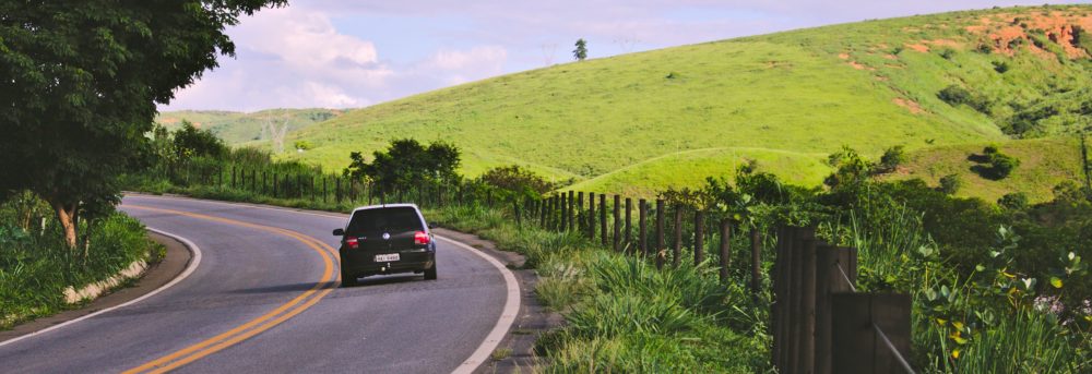 Single black car driving on two lane road. To the right is grass, a fence and up ahead a bright green hill. The sky is cloudy