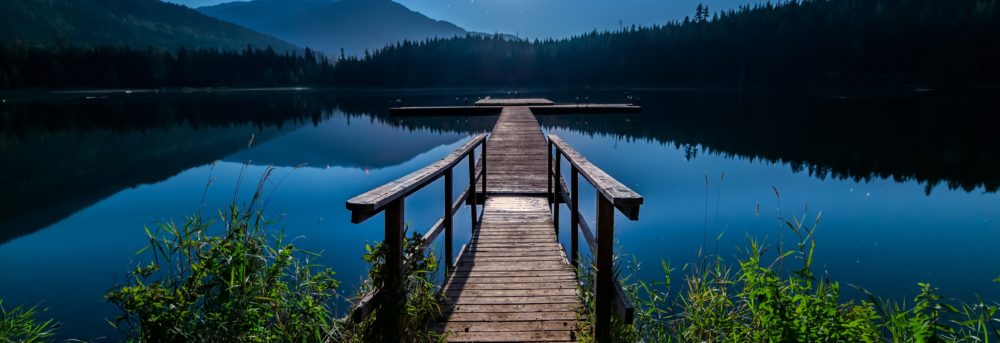 Wooden bridge with handrails extending into blue water with greenery on both sides. The moon is full, hanging above a mountain range on the other side of the water