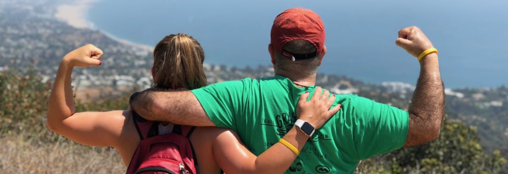 Molly and Dan standing with their backs to the camera overlooking the ocean near Santa Monica. Molly is making a muscle with her left arm and carrying a red Nike Stanford backpack. Dan is wearing a green shirt and red baseball cap and is making a muscle with his right arm.