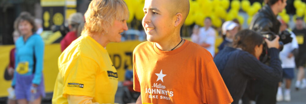 Jimmy in Austin in 2008 after high dose chemo receiving a yellow rose. He's wearing an orange Mellow Johnny's t-shirt, a race bib with the number 28 on it. He's bald. Behind him are photographers, a volunteer in a yellow LIVESTRONG shirt and photographers