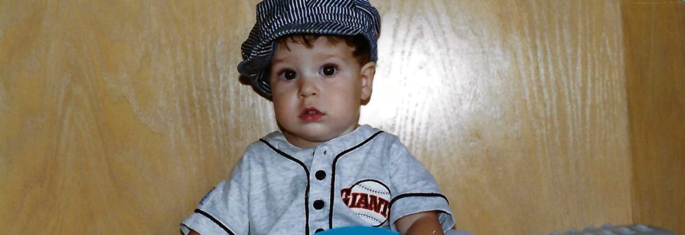 Jimmy at age 1 wearing a striped train engineer cap, an SF Giants light gray onesie with Giants on his left breast sitting with trains from Thomas the Tank Engine