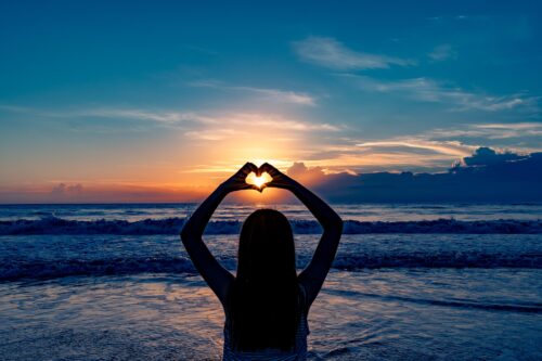Woman standing in front of the ocean at sunset. Her arms are above her head, her hands making the shape of a heart that frames the setting sun