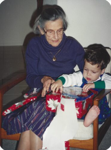Jimmy age 1.5 opening a christmas present. He's sitting next to Mom who is wearing a navy blue sweater, wool skirt and glasses. 