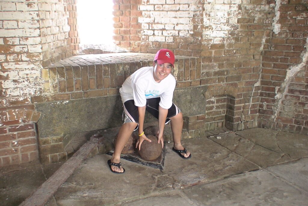 Jimmy squating down with his hands on an antique cannonball on a chain. He's wearing a red Red Dox hat, a white t-shirt, black shorts with white piping and flip flops
