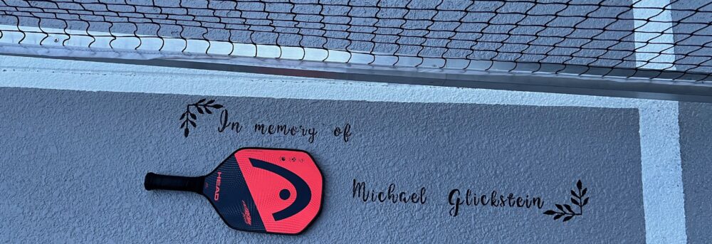 A pickleball racquet in its red and black case laying next to the net with the words "In memory of Michael Glickstein" written on the ground