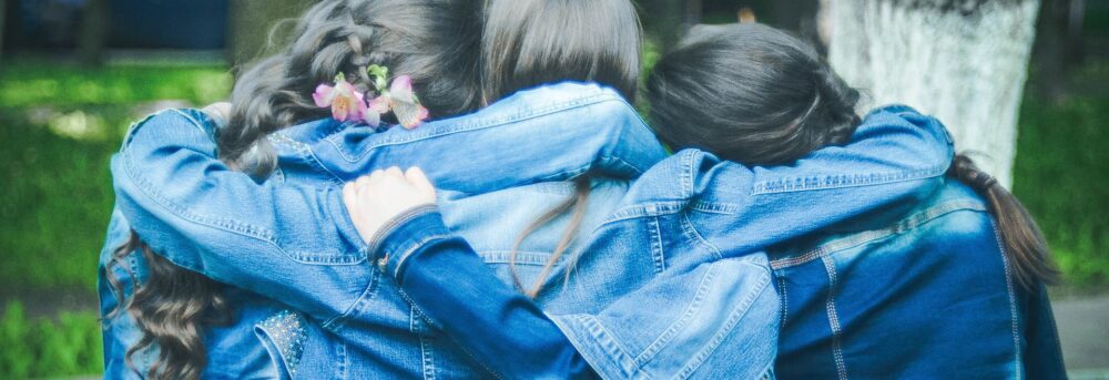 Three young friends with their arms around each other. They're all wearing jean jackets and their backs are to the camera
