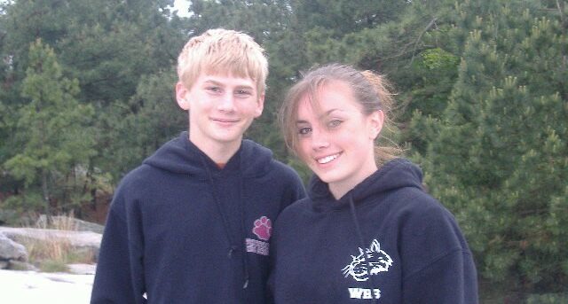 TJ and Jessica standing next to each other. They are both wearing navy sweatshirts with logos on the left side. TJ has short blond hair. Jessica has darker blond hair that's pulled back into a pony tail