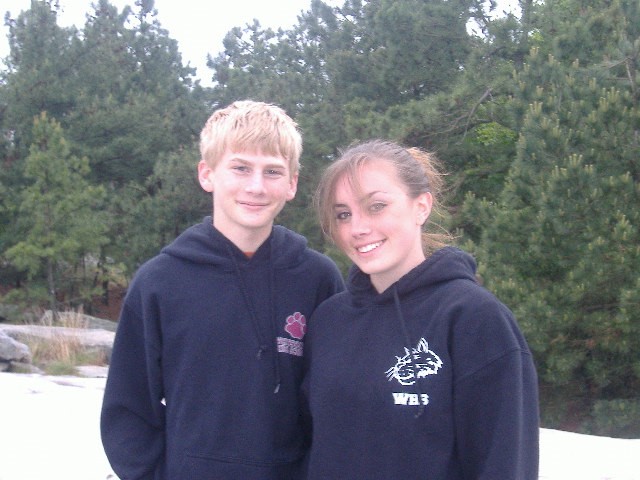 TJ and Jessica standing next to each other. They are both wearing navy sweatshirts with logos on the left side. TJ has short blond hair. Jessica has darker blond hair that's pulled back into a pony tail