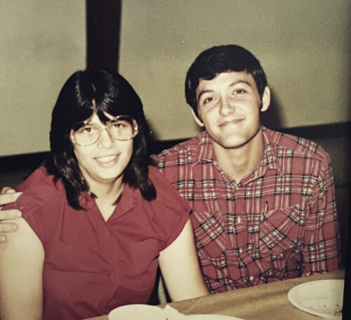 Paula and Forrest as young adults. Paula is wearing a short sleeve maroon button shirt with a color and Forrest is wearing a red and black plaid button shirt. Paula is wearing glasses; they both have dark hair