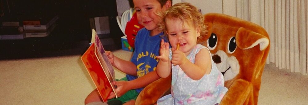 Jimmy and Molly sitting next to each other in small animal chairs. Jimmy is reading to Molly wearing a blue t-shirt. Molly is wearing a sleeveless dress and holding a pretzel in her hand.